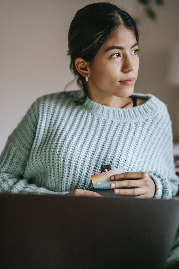 Young dreamy ethnic female buyer with debit card and netbook looking away in house room